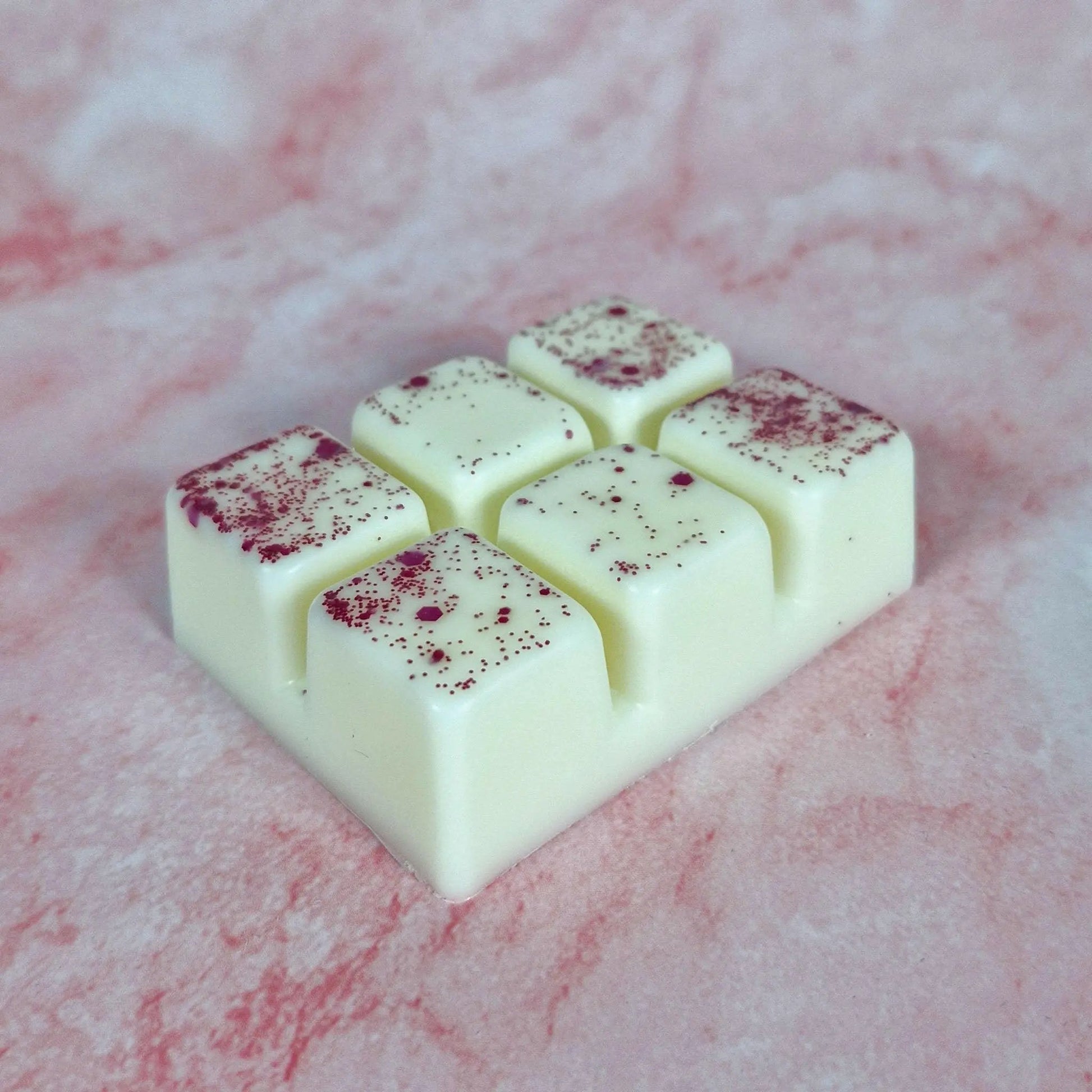 Close-up of Rose Petals wax melt, six off-white cubes with red glitter, on a pink textured background.