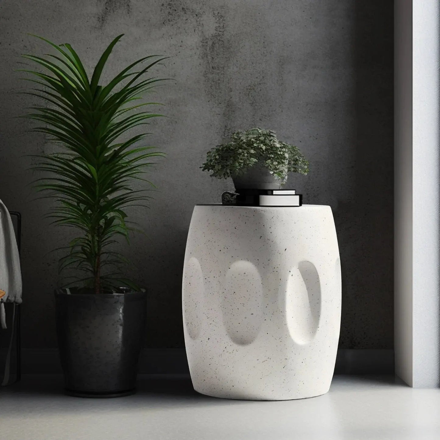 Modern round lightweight concrete side table with textured indents, next to a tall plant in a dark room.