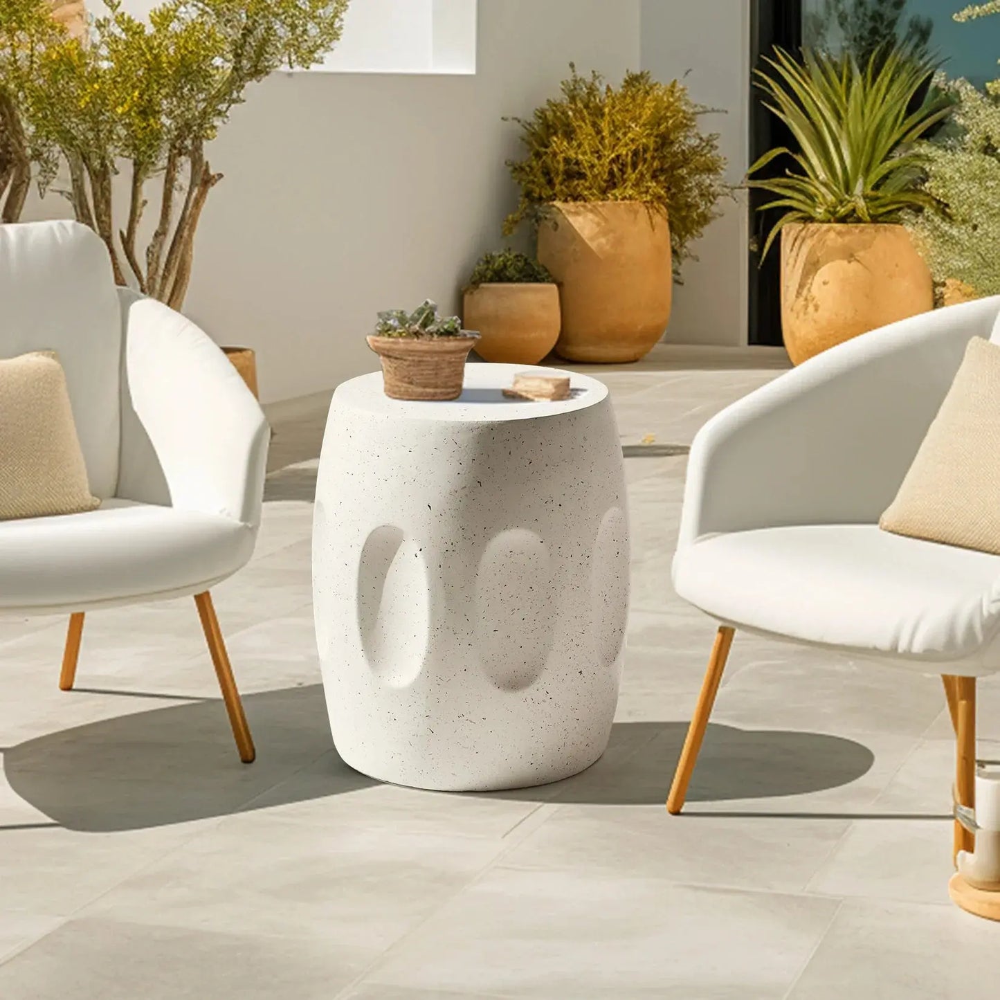 Modern round lightweight concrete side table with textured indents, next to two white patio chairs on a tiled outdoor space.