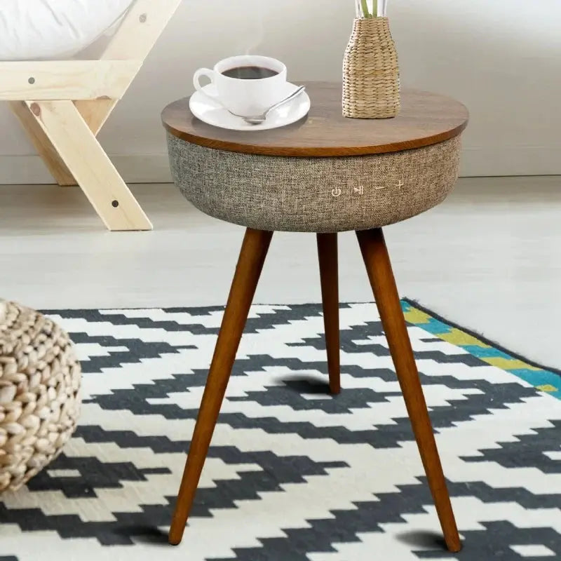 Round wooden smart coffee table with grey speaker fabric, controls, and three legs, holding a coffee cup.