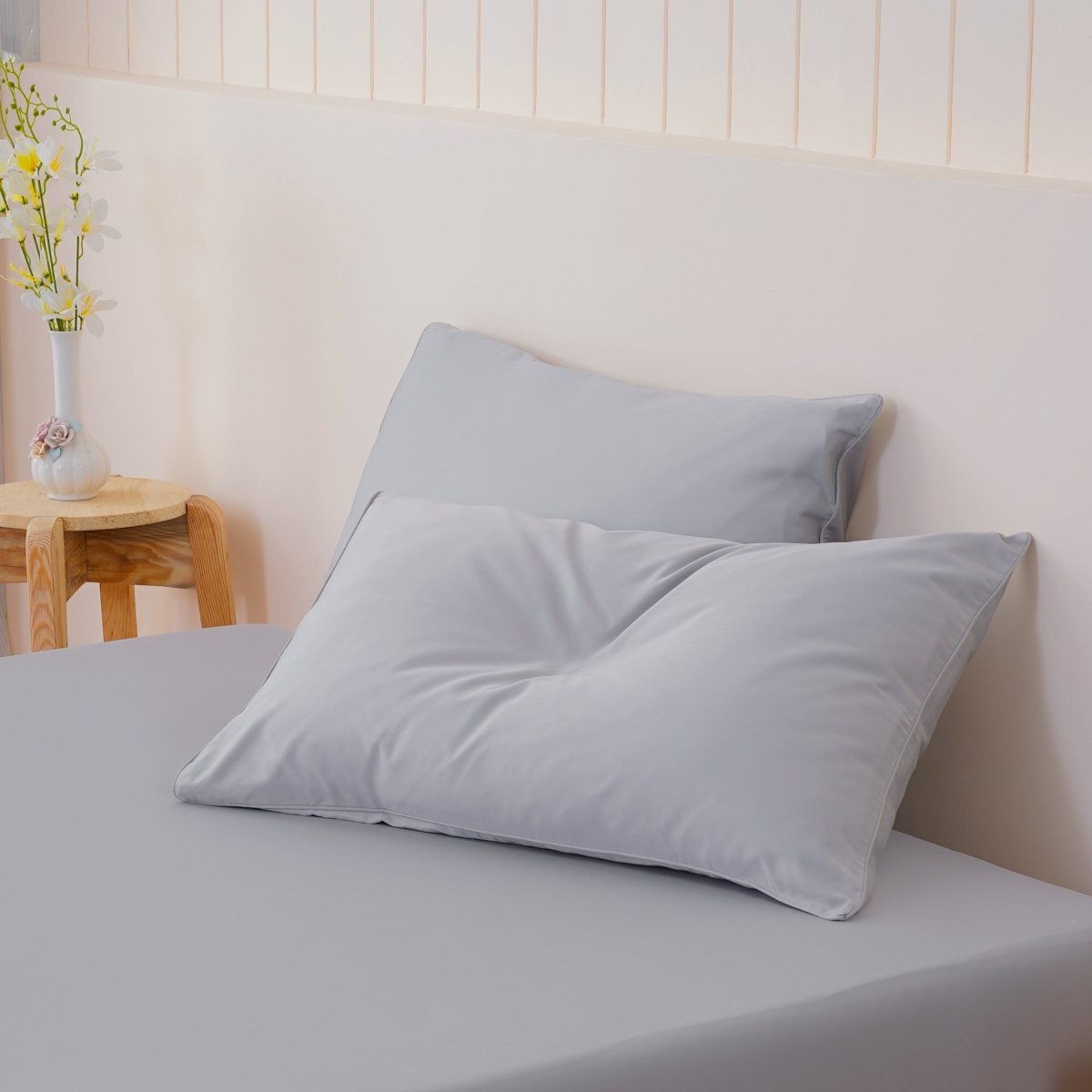 Gray pillows on a bed with a wooden side table and flowers in the background