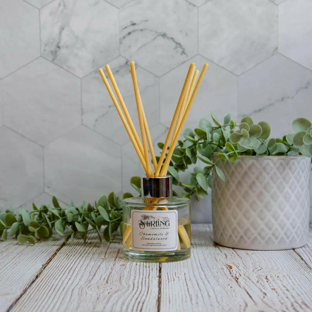 Chamomile & Sandalwood 100ml diffuser with reeds on a wooden surface, next to a plant, against a marble wall.