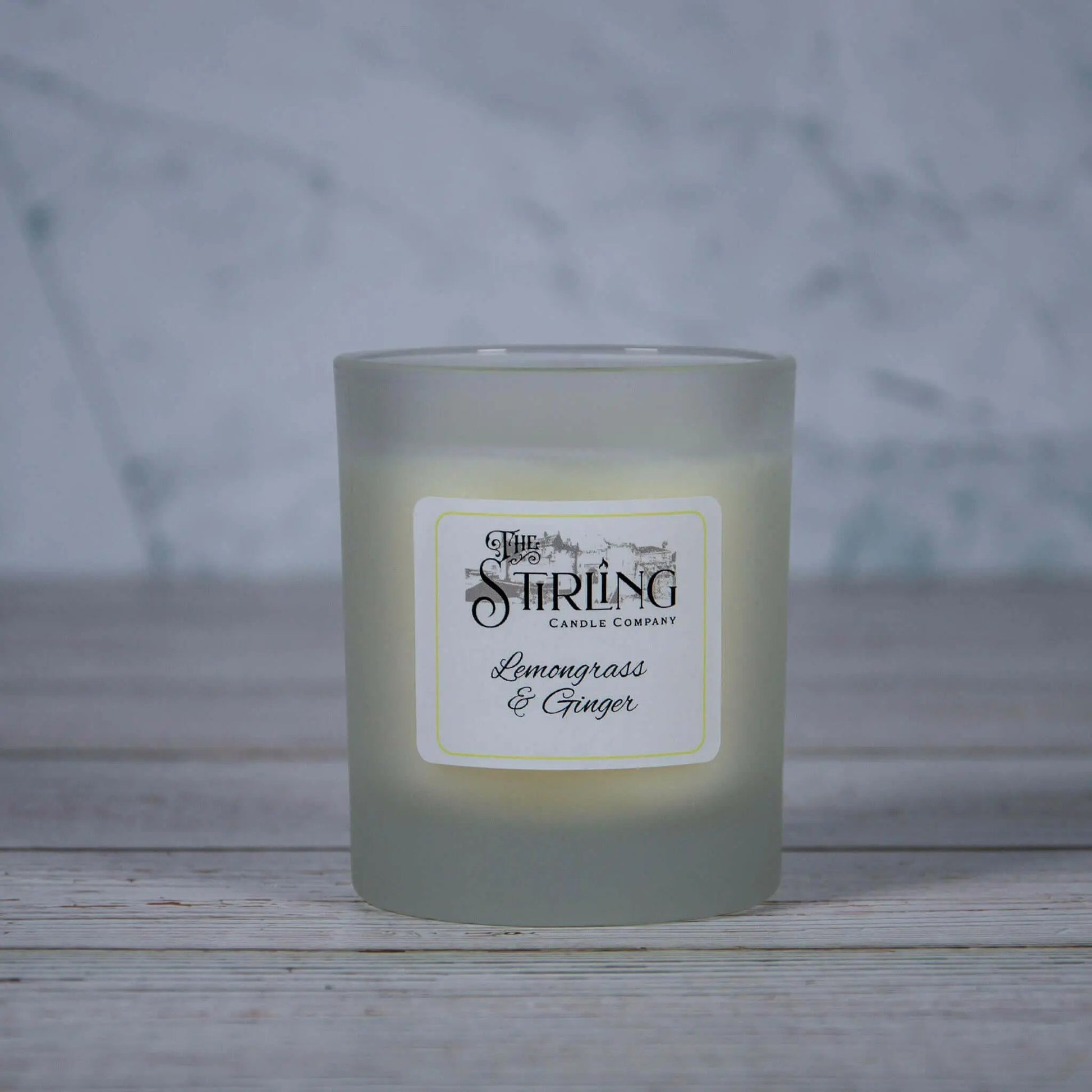The Stirling Candle Company Lemongrass & Ginger medium candle in a frosted glass jar, on a white wooden surface.