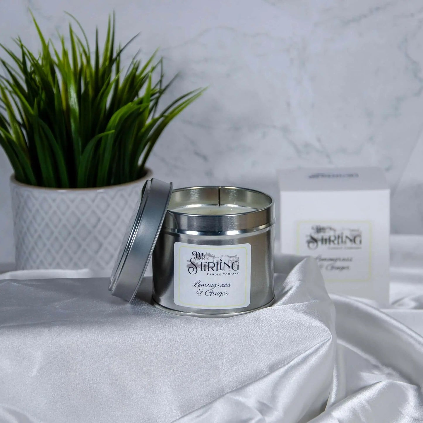 Lemongrass & Ginger travel tin candle with wooden wick, partially open, on white fabric with plant and box in background.