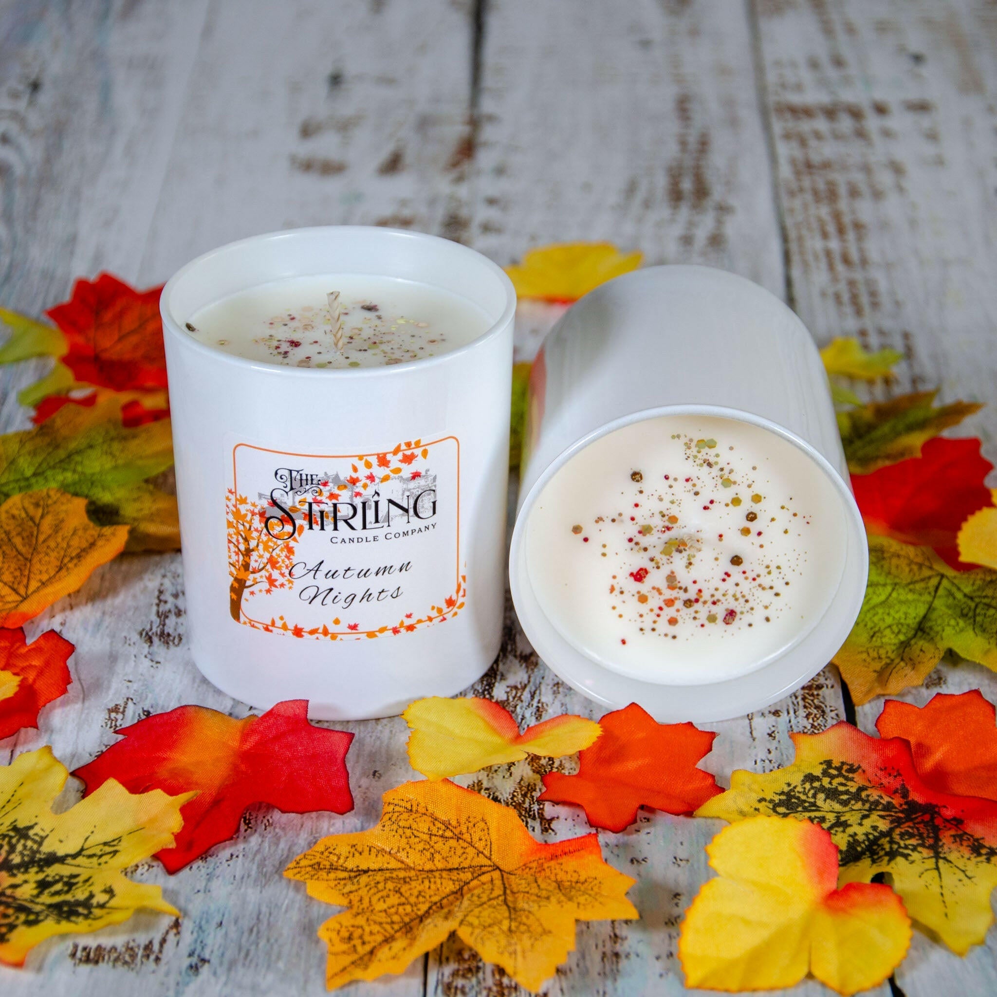 Two Autumn Nights large candles, white with autumn-themed labels, on a rustic wooden background with fall leaves.