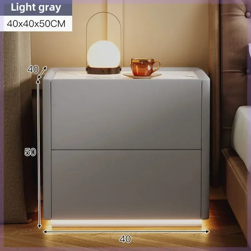 Light gray modern nightstand with two drawers, illuminated base, and a white marble top. Features a lamp and teacup.