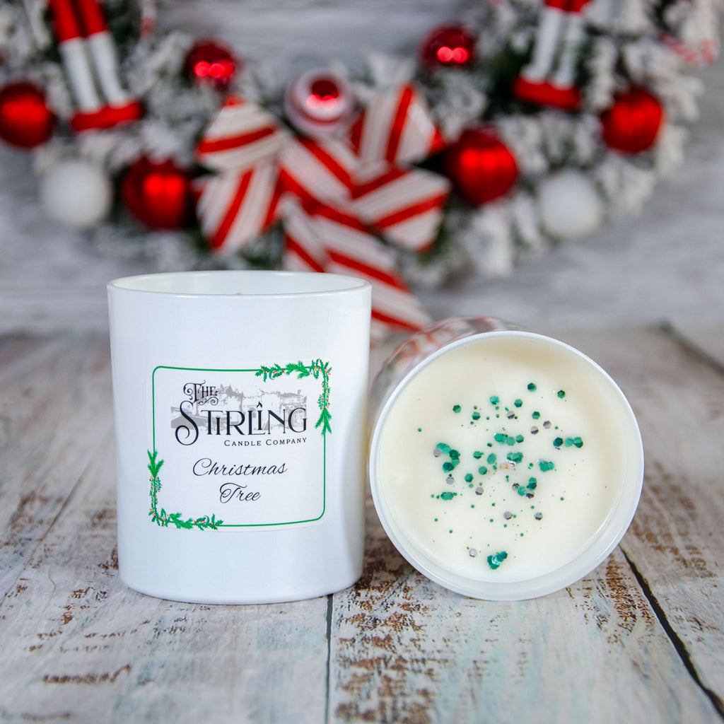 Large Christmas Tree candle with white label, beside open candle with green glitter on white wax. Festive wreath in background.