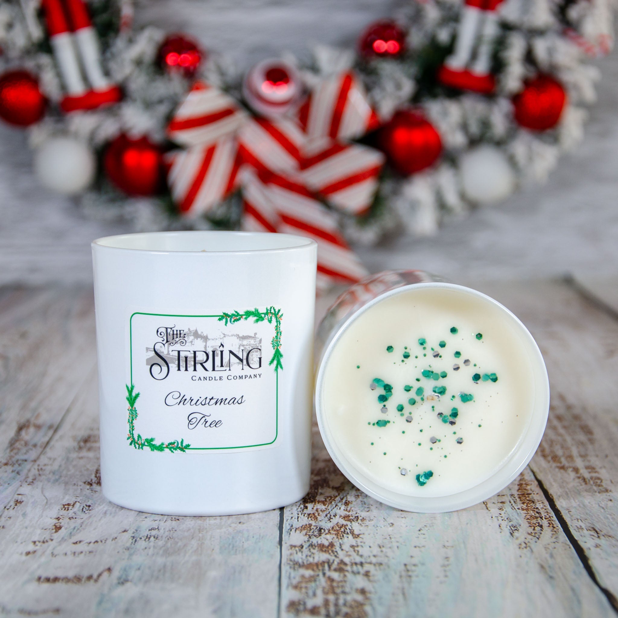 Large Christmas Tree candle with white label, beside open candle with green glitter on white wax. Festive wreath in background.