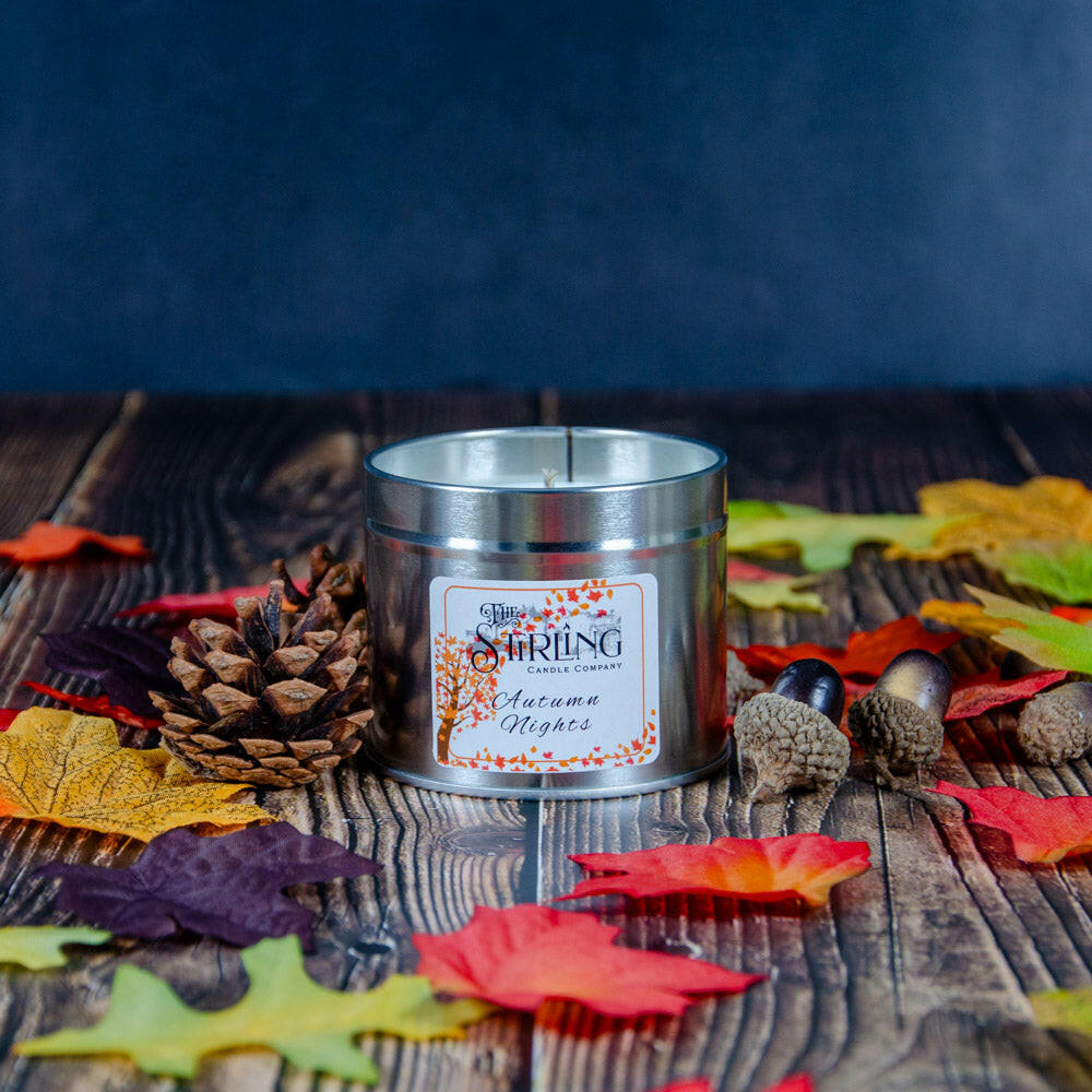 Autumn Nights Travel Tin Candle by The Stirling Candle Company, surrounded by autumn leaves, pinecone, and acorns on wood.