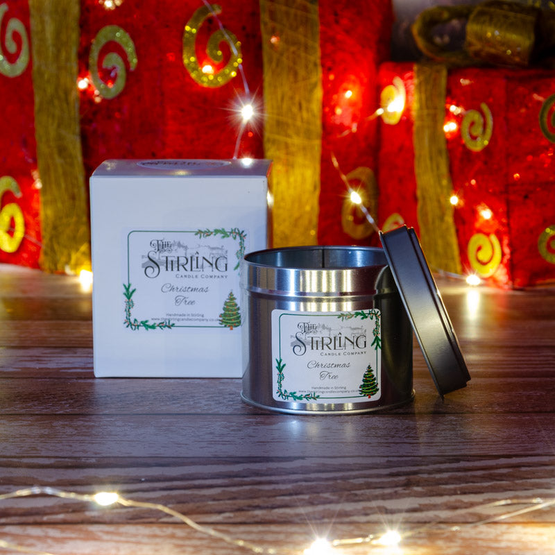 Christmas Tree travel tin candle with cotton wick next to its white box, on wood with festive lights.