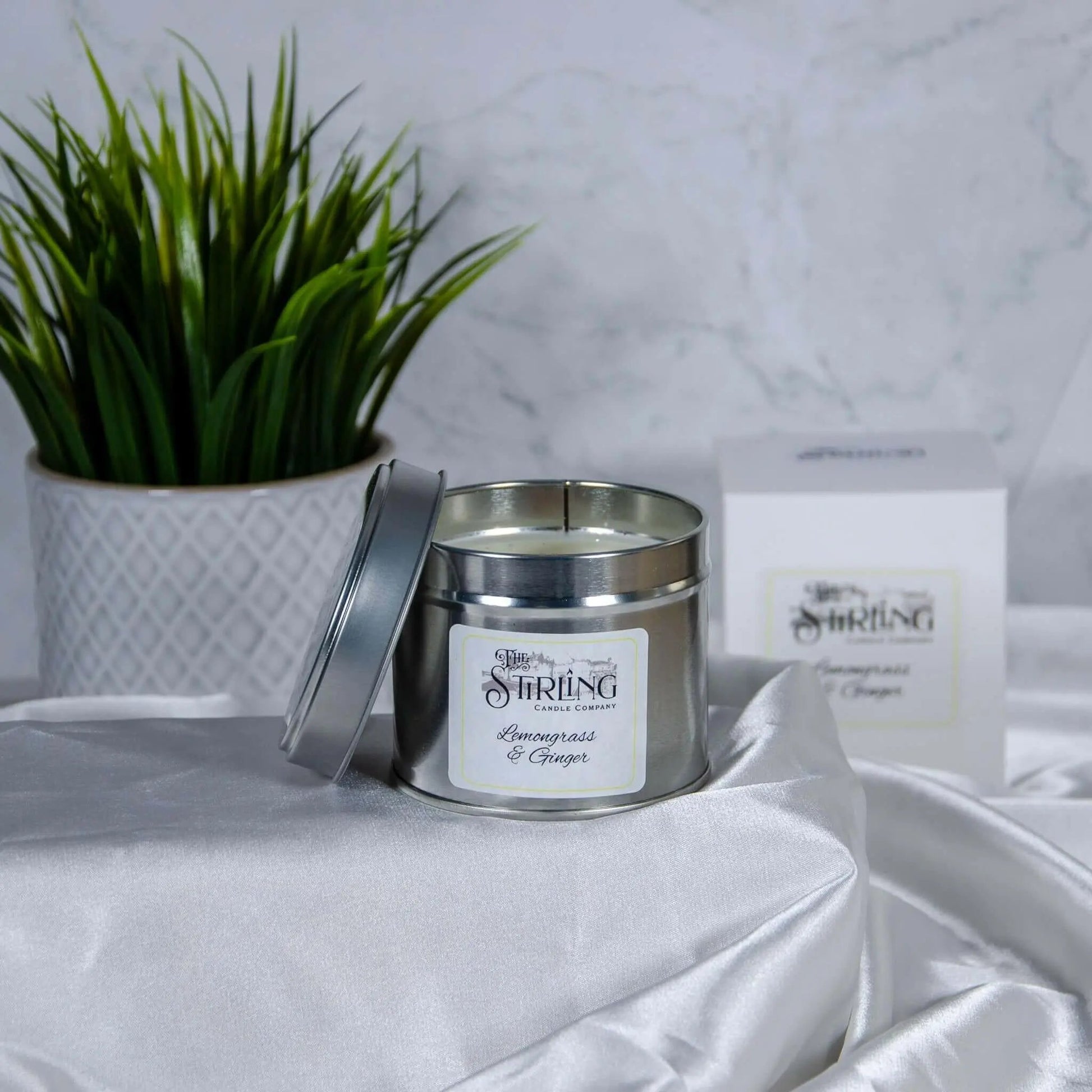 Lemongrass & Ginger travel tin candle with wooden wick, partially open, on white fabric with plant and box in background.