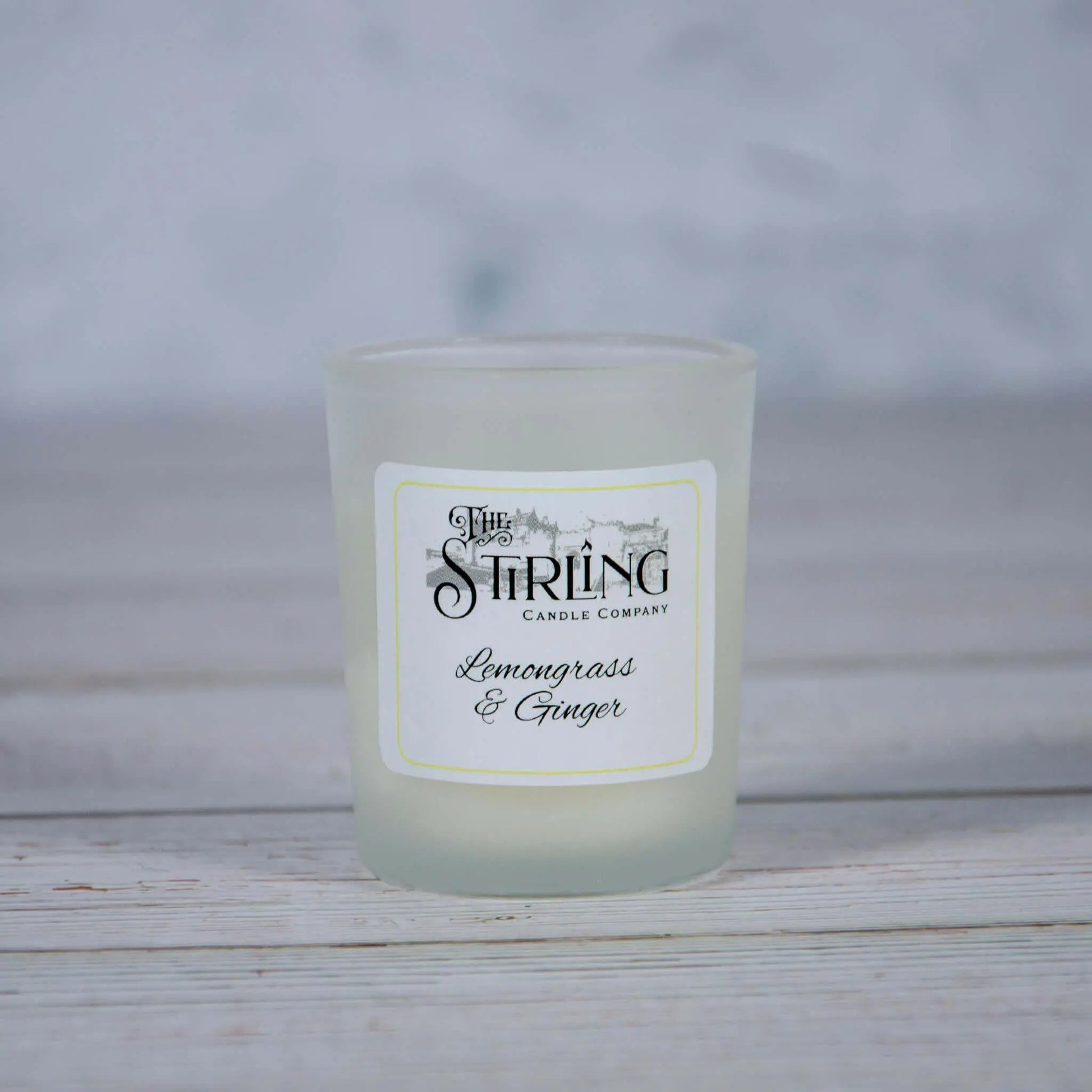 Small frosted glass Lemongrass & Ginger scented candle by The Stirling Candle Company on a light wooden surface.